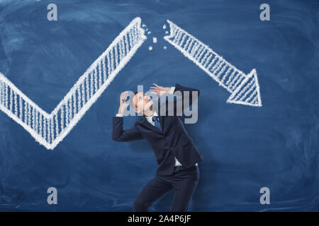Businessman cowering on blue blackboard background with chalk drawing of white statistic arrow broken in half. Stock Photo