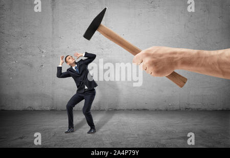 Scared businessman full-height in a desperate pose and a giant hand with a hammer threting him on the gray background Stock Photo
