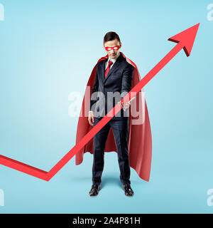 A businessman in a red flowing cape trying to keep the upward looking red arrow with his hand. Stock Photo