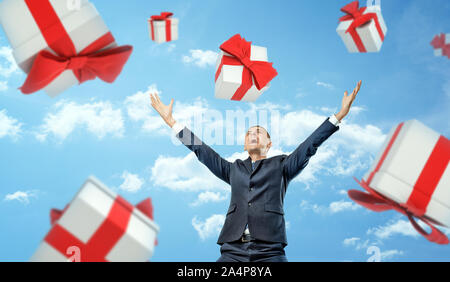 A happy businessman standing with hands raised in victory motion under a rain of gift boxes falling on him. Stock Photo