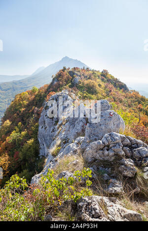 Distant pointy peak and a rocky mountain path covered by trees and bushes with vivid, autumn colors of the leaves Stock Photo