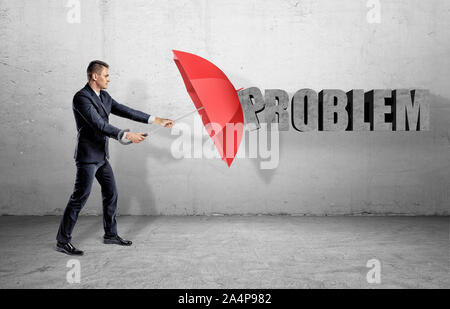 A businessman hiding behind a red open umbrella from a word 'Problem' on the wall. Stock Photo