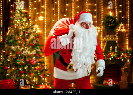 Santa Claus with a big sack of gifts at his room at home near Christmas tree. Getting ready to go Stock Photo