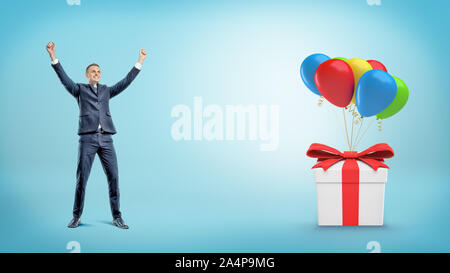A happy businessman with hands raised up standing behind a gift box tied to many balloons. Stock Photo