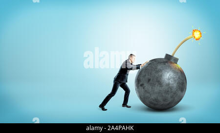 A tiny businessman in side view pushing at a giant round bomb with a lit fuse on blue background. Stock Photo