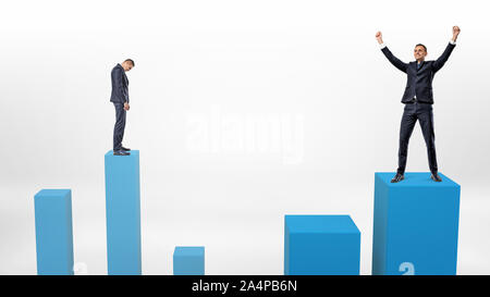 A sad businessman prepares to step down on a low column and a winner beside him who just stepped up on a higher one. Stock Photo