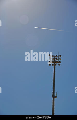 Airplane flying high in the sky, leaving a contrail behind. Lighting pylon of a basketball court in the foreground. Stock Photo