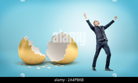 A victorious businessman stands near a large broken golden eggshell on blue background. Stock Photo