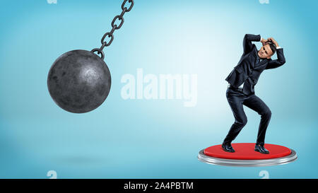 A scared cowering businessman stands on a huge push button and hides his head from a wrecking ball. Stock Photo