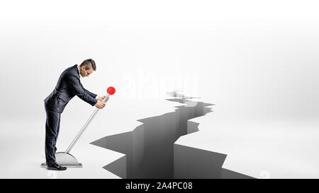 A businessman turns a large metal lever while standing beside a long earthquake rift on white background. Stock Photo