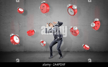A businessman on a concrete background looks up and hides from many red retro alarm clocks falling down. Stock Photo