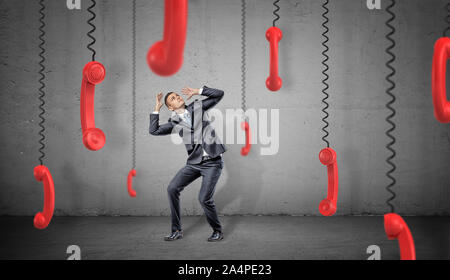 A scared businessman on concrete background hides from many red retro phone receivers hanging down on their cords. Stock Photo
