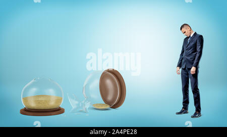A sad businessman with his head low stands near a giant broken hourglass on blue background. Stock Photo