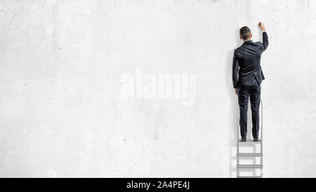 A businessman stands on a step ladder in a back view and draws on an empty white wall. Stock Photo