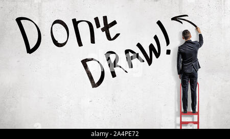 A businessman stands on a red ladder and makes a writing Don't Draw on a black wall. Stock Photo