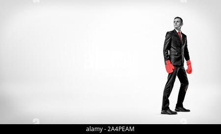 A businessman stands in half turn on a while background wearing a suit and red boxing gloves. Stock Photo