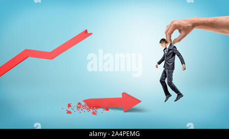 A small businessman is lifted up by a giant human hand on a blue background over a red broken statistic arrow. Stock Photo