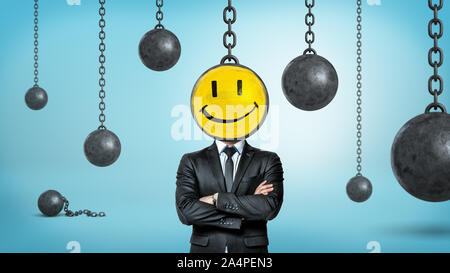 A businessman with folded arms stands surrounded by wrecking balls an one hiding his face with a smiley emoticon. Stock Photo