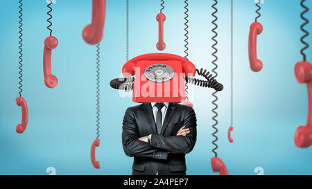 A businessman with folded arms stands surrounded by phone receivers and with one red retro phone placed on his head. Stock Photo