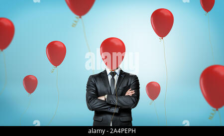 A businessman in front view with crossed arms stands surrounded by many red party balloons with one hiding his face. Stock Photo
