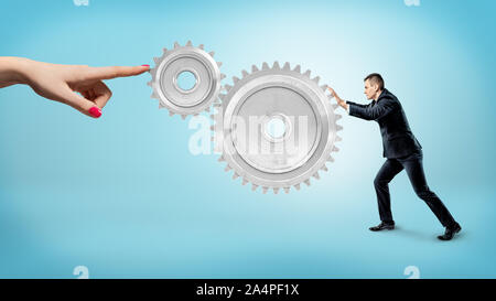 A small businessman pushes at a large gear interlocked with a small one while a female finger stops their movement. Stock Photo