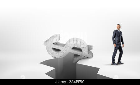 A businessman stands in half turn near a large earthquake crack on a white surface with a giant concrete dollar sign falling inside. Stock Photo