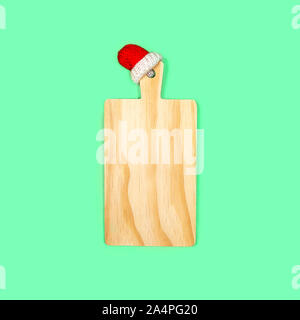 Christmas New Year concept with cherry wooden kitchen cutting board in Santa Claus red hat on trendy mint green blue color background. Flat lay, top view, copy space. Stock Photo