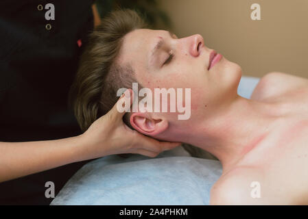The masseur does a facial massage to a young guy. A man is massaged face on a massage table. Stock Photo
