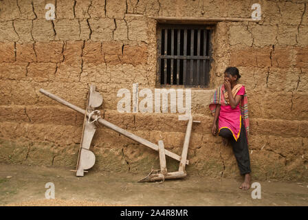 A girl from the Santal community leans beside a traditional wooden plow, against the wall of a mud home, in Joypurhat, Rajshahi, Bangladesh. May 20, 2011. .The Santals are an ethnic minority community living in different districts of Rajshahi division in Bangladesh.  Most do not own land and work as daily paid labourers for very low wages. Much of the advancements in technology have bypassed these isolated communities where males dominate and women shoulder the burden of farming and carrying out other livelihoods..Santals are also the largest ethnic group in India where they have settlements i Stock Photo