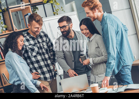 Startupers working together at office standing bearded man pointing at laptop explaining project cheerful while others looking at screen Stock Photo