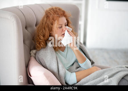 Woman sitting in armchair and sneezing because of flu