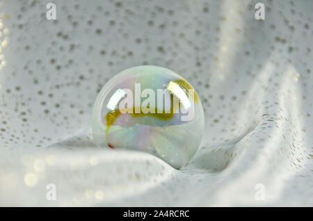 A large polished and shimmering marble, bullet of glass, in colour of nacre lying on a ground of white and gold sparkling texture Stock Photo