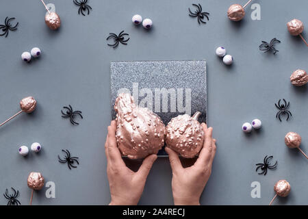 Hands trying to fit two pumpkins on decorative mirror. Creative Halloween flat lay background in grey, pink and black with chocolate eyes and spiders. Stock Photo
