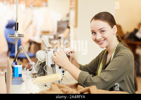 Portrait of young tailor sitting and smiling at camera while sewing on sewing machine in atelier Stock Photo