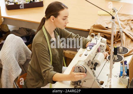 Serious young woman sitting and sewing using sewing machine in atelier Stock Photo