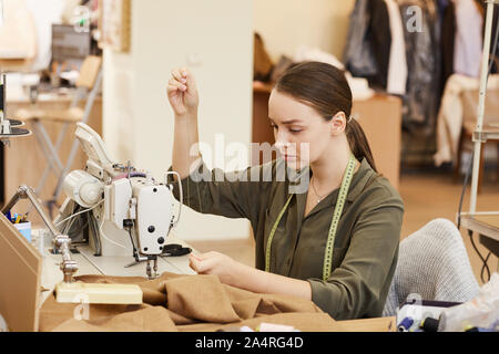 Young seamstress sitting at the table in front of sewing machine and using needle and thread for sewing clothes Stock Photo