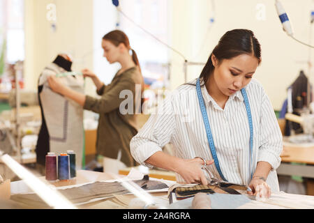 Concentrated Asian designer working with scissors she cutting fabric at the table while other designer working with mannequin in the background Stock Photo