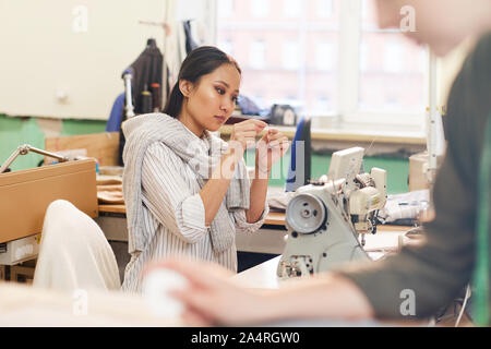 Serious Asian tailor threading the needle while sitting at the table in front of sewing machine in workshop Stock Photo