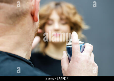 Hairdresser is using hairspray on client's hair at salon. Closeup of hairdresser's hands are using hairspray on client's hair on gray background. Stock Photo