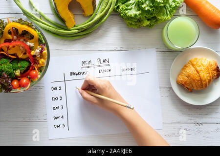 Calories control, meal plan, food diet and weight loss concept. top view of hand filling meal plan on weekly table with salad and fresh vegetable on d Stock Photo