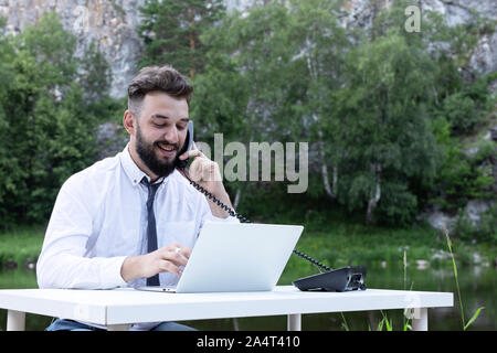 Office worker guy at desk using desktop computer laptop and phone at work, smiling, looking at screen. Outdoors Stock Photo