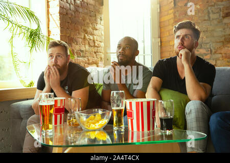 Group of friends watching game on TV at home. Sport fans spending time and having fun together. Emotional, expressive, exciting game. Cheer for favorite football or soccer team, friendship, weekend. Stock Photo