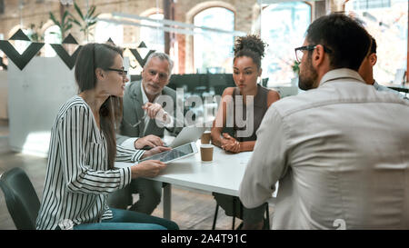 Using digital technologies. Young business woman holding digital tablet and discussing something with colleagues while sitting at the office table tog Stock Photo