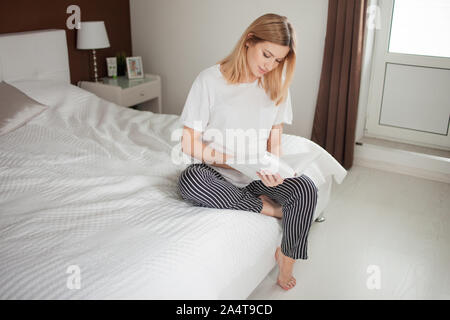 The expectant mother sits on the bed with one leg dangling, and enthusiastically reads a book and strokes her tummy. A pregnant woman reads literature Stock Photo