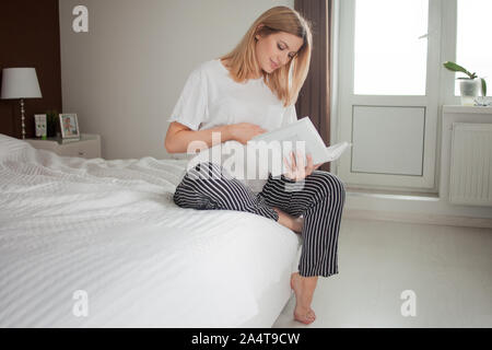 The expectant mother sits on the bed with one leg dangling, and enthusiastically reads a book and strokes her tummy. A pregnant woman reads literature Stock Photo