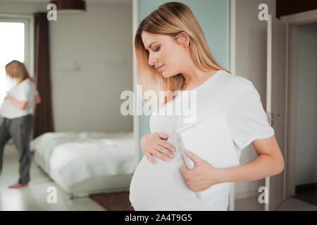 Portrait of a pregnant woman stroking her tummy, with a white book under her arm against the background of reflection in the mirror. Side view. Stock Photo