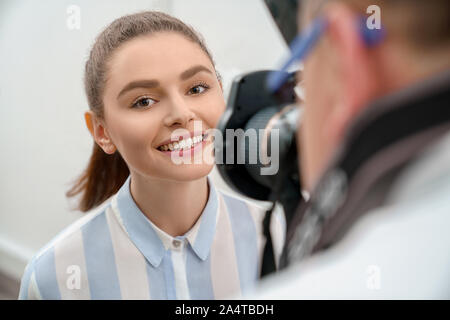 Pretty woman looking at camera, smiling. Charming, adorable model with brilliant smile posing for photographer and showing white teeth. Man holding camera, taking photo. Stock Photo