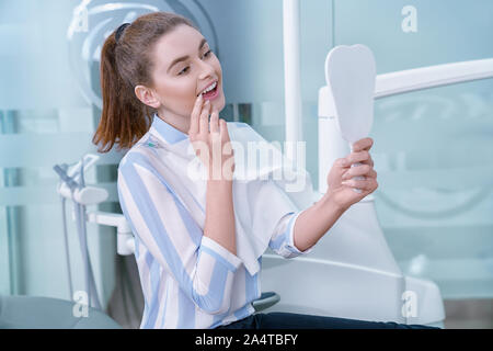 Happy woman satisfied with professional private clinic of dentistry. Charming and adorable woman looking at mirror with opened mouth, smiling. Client holding tooth shaped mirror. Stock Photo