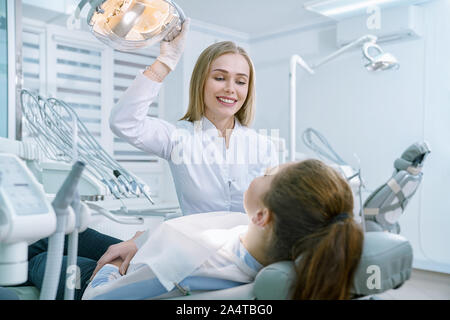 Doctor working with patient in private clinic. Professional doctor wearing in white medical coat and gloves. Charming blonde smiling, looking at patient. Client lying in dentist chair. Stock Photo