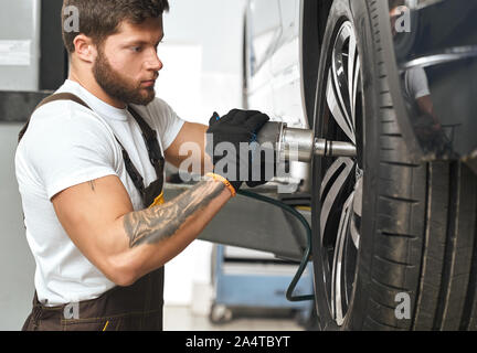 Bearded, brutal mechanic fixing hubcap of auto wheel, using equipment. Muscular man in white t shirt with tattoo on hand wearing in black gloves, working in autoservice. Stock Photo